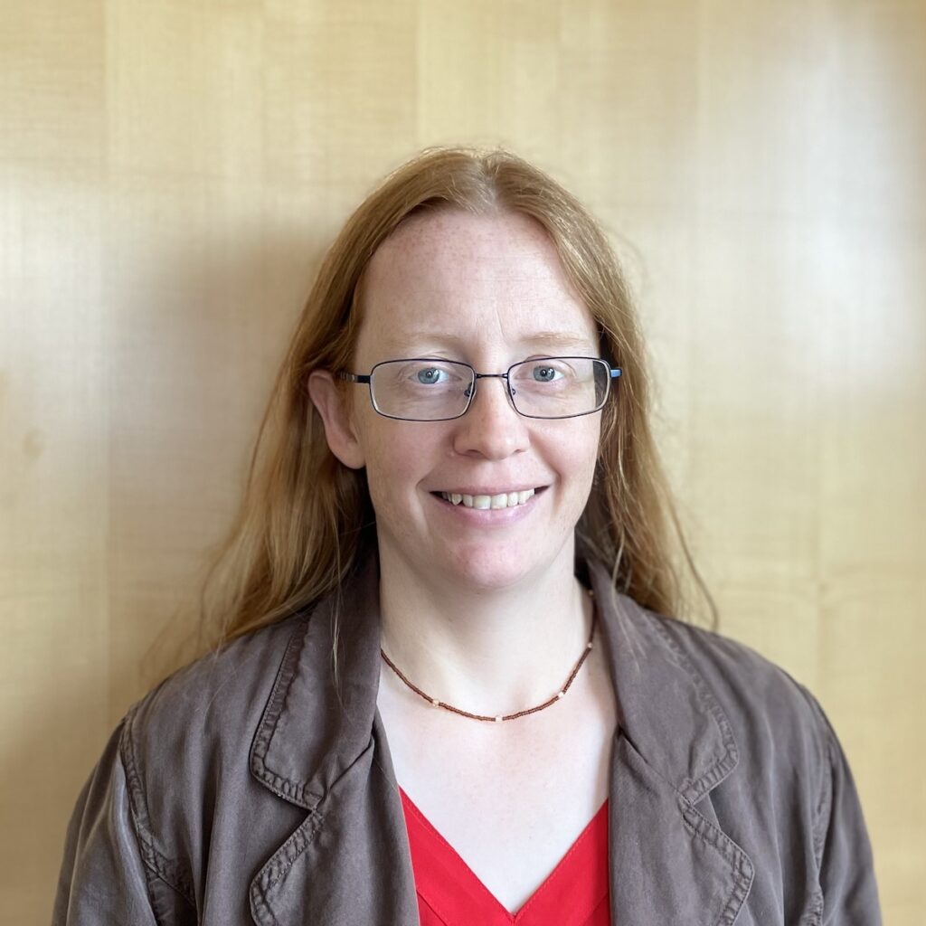 headshot2023 Headshot of a white woman with red hair and glasses, wearing a red shirt and brown jacket, standing in front of a wooden door.