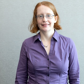 A White woman with red hair and glasses wearing a purple button-down shirt