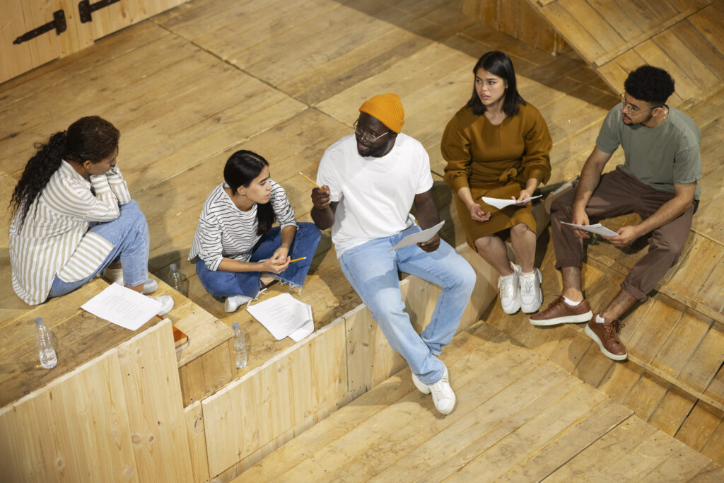 Five ethnically diverse people sit on the edge of a wooden stage with scripts. A Black man in jeans, a white t-shirt, and an orange beanie gestures with a pencil while giving instructions to two young people, while another young woman and a young man look on.