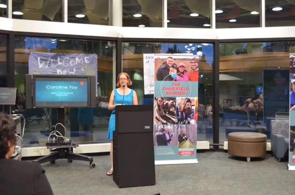 A White woman with red hair wearing a bright blue dress stands speaks from behind a podium. There is a large banner reading "Ohio Field School" to her left and a screen reading "Caroline Toy" to her right.