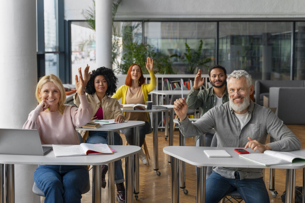 A diverse group of adults sitting at desks in a conference or classroom, all with one hand raised.